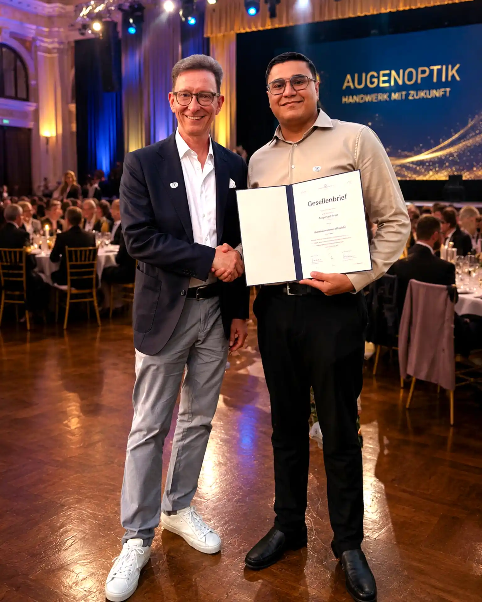 Gratulation zur Gesellenprüfung im Augenoptiker-Handwerk. Zwei Personen mit Urkunde auf einer Veranstaltung.